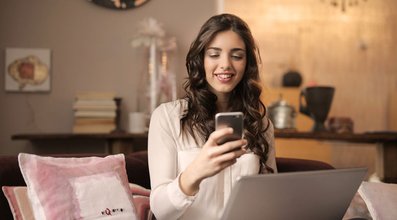 Mujer mirando su celular y sonriendo frente a una computadora