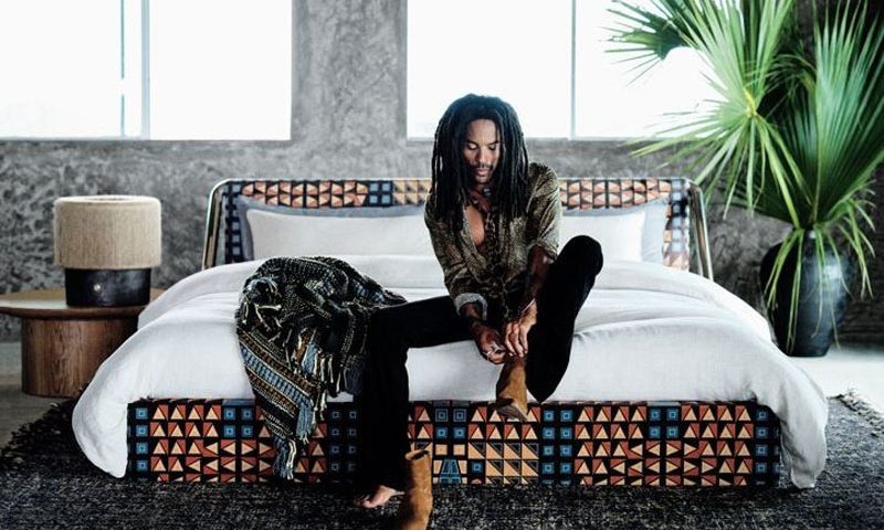 Lenny Kravitz cambiándose los zapatos en el borde de una cama de CB2 x Kravitz