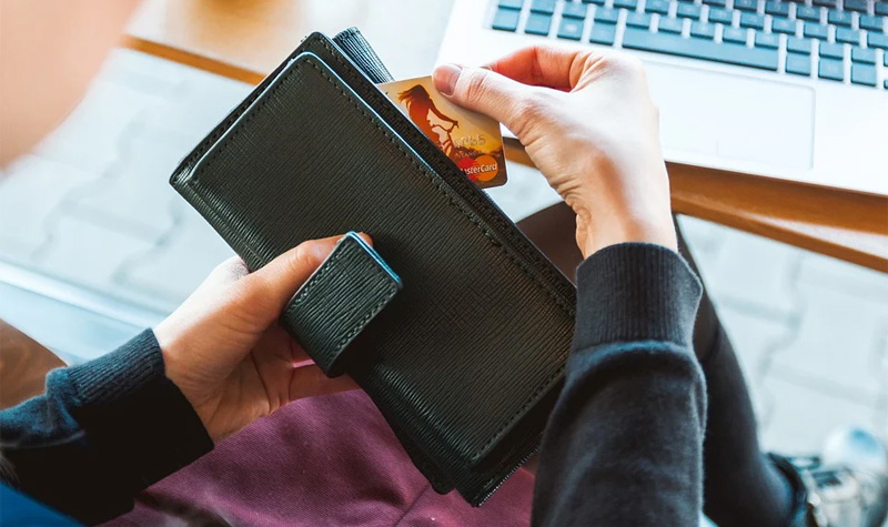 Una persona saca su tarjeta de crédito de la billetera mientras está sentada frente a su notebook