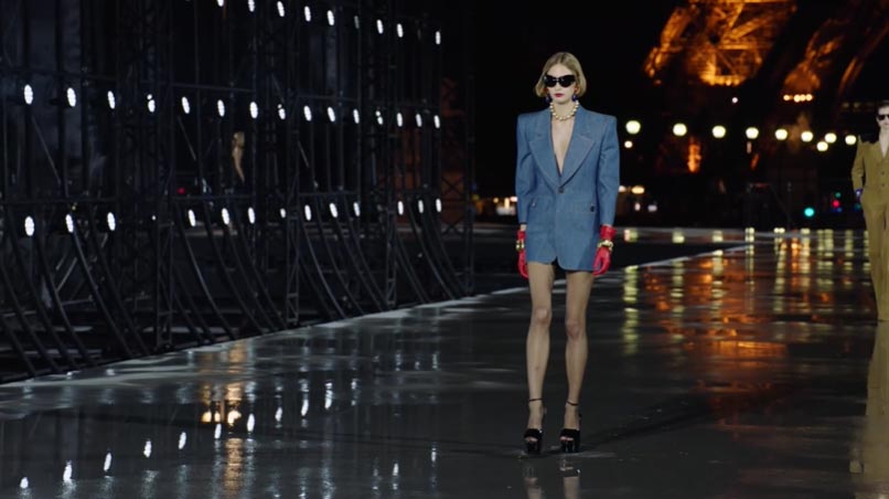 Modelo de Saint Laurent en traje ochentoso de denim con guantes rojos y gafas oscuras desfila en París