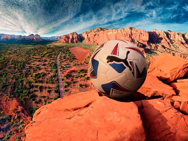 pelota oficial de la CONMEBOL Copa América 2024, Puma, apoyada en la coordillera sobre tierra roja al borde de una montaña. Detrás se ve el horizonte