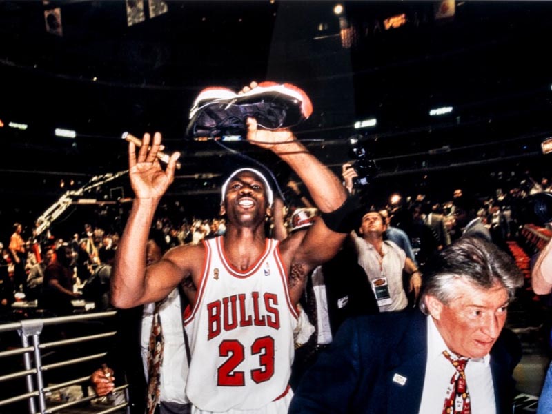 Michael Jordan levantando una zapatilla en alto vestido con la camiseta de los Chicago Bulls en un estadio de basket